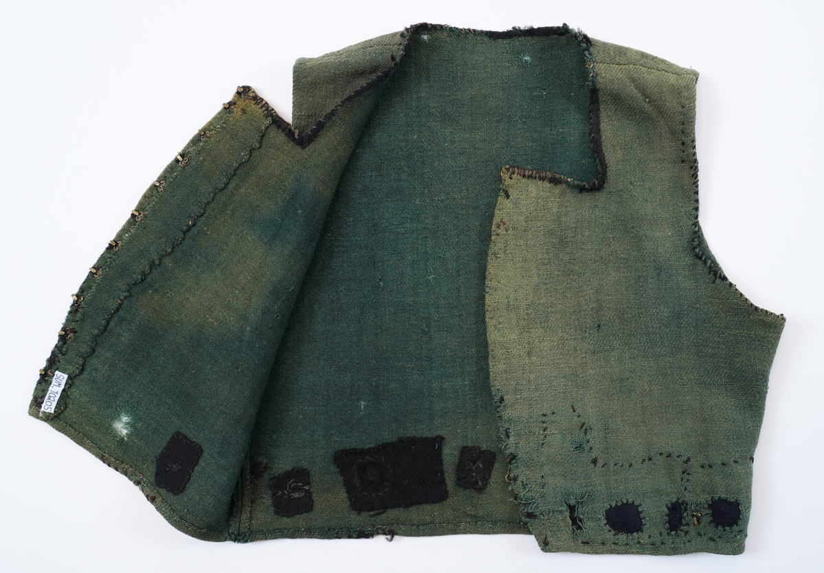 Livstykke som er overdel til kvinnedrakt. Livstykke i grønn ull med firskoren halsopning. Skal festast heilt ihop framme med 11 sett hekter og hemper. Hekter og hemper rundt livet til feste for skjørt. Rundt ermeopningane har det vore enkel fald, seinare er dette stadvis forsterka med overkast av grov tråd. Det same er tilfellet rundt halsopningen der det opphavleg har vore svart ullband. Livstykket er reparert med svarte og blå bøter i nedkant. Framkanten på livet er runda for å lage plass til bysten.