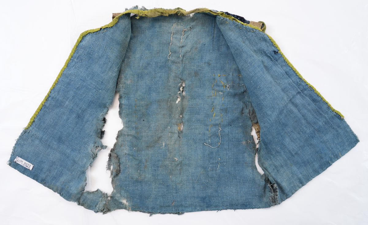 Opplut i blå plysj med blått linfôr. Rund halsringing og litt skrådd opning framme. Ikkje skuldersaum, men eit insnitt på skuldra mot ermopningen. Framkant og hals er kvarda med eit gulgrønt lerretsvove silkeband, og ermopning er kvarda med grønt silkeband med lys jarekant. Ermopning på bakstykke og eit stykke ned på framstykke er prydd med gylte sølvborder. Ryggen har ei bord midt på  og i to motståande vinklar på kvar side av midtborda.
Lause trådar i ermopningen tyder på at det har vore ermer i oppluten.
Det er 2 skøyter på framstykket ein 23 cm ned frå skulder og ein 39 cm ned.