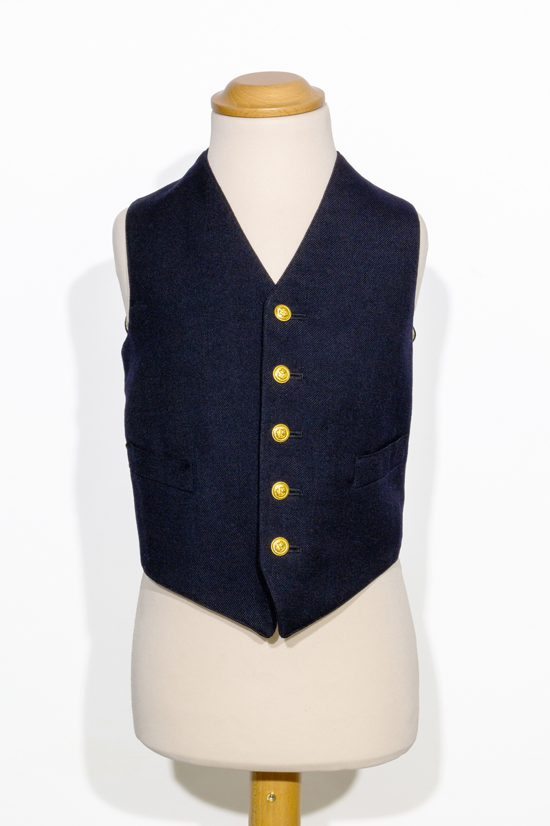Uniformsväst för barn i mörkblått ylletyg. Västen har två fickor, en på vardera sidan om den främre knäppningen. Västen har fem stycken gulfärgade knappar med motiv av ankare. Västens bakstycke är i svart ylletyg och försedd med ett spänne. Västen är fodrad med ett randigt tyg med bruna och blå ränder på oblekt bas. 