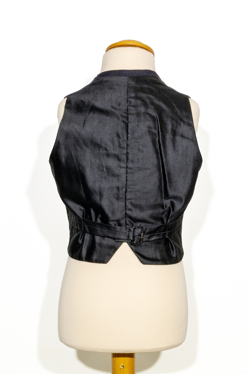 Uniformsväst för barn i mörkblått ylletyg. Västen har två fickor, en på vardera sidan om den främre knäppningen. Västen har fem stycken gulfärgade knappar med motiv av ankare. Västens bakstycke är i svart ylletyg och försedd med ett spänne. Västen är fodrad med ett randigt tyg med bruna och blå ränder på oblekt bas. 