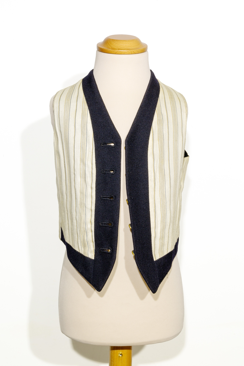 Uniformsväst för barn i mörkblått ylletyg. Västen har två fickor, en på vardera sidan om den främre knäppningen. Västen har fem stycken gulfärgade knappar med motiv av ankare. Västens bakstycke är i svart ylletyg och försedd med ett spänne. Västen är fodrad med ett randigt tyg med bruna och blå ränder på oblekt bas. 