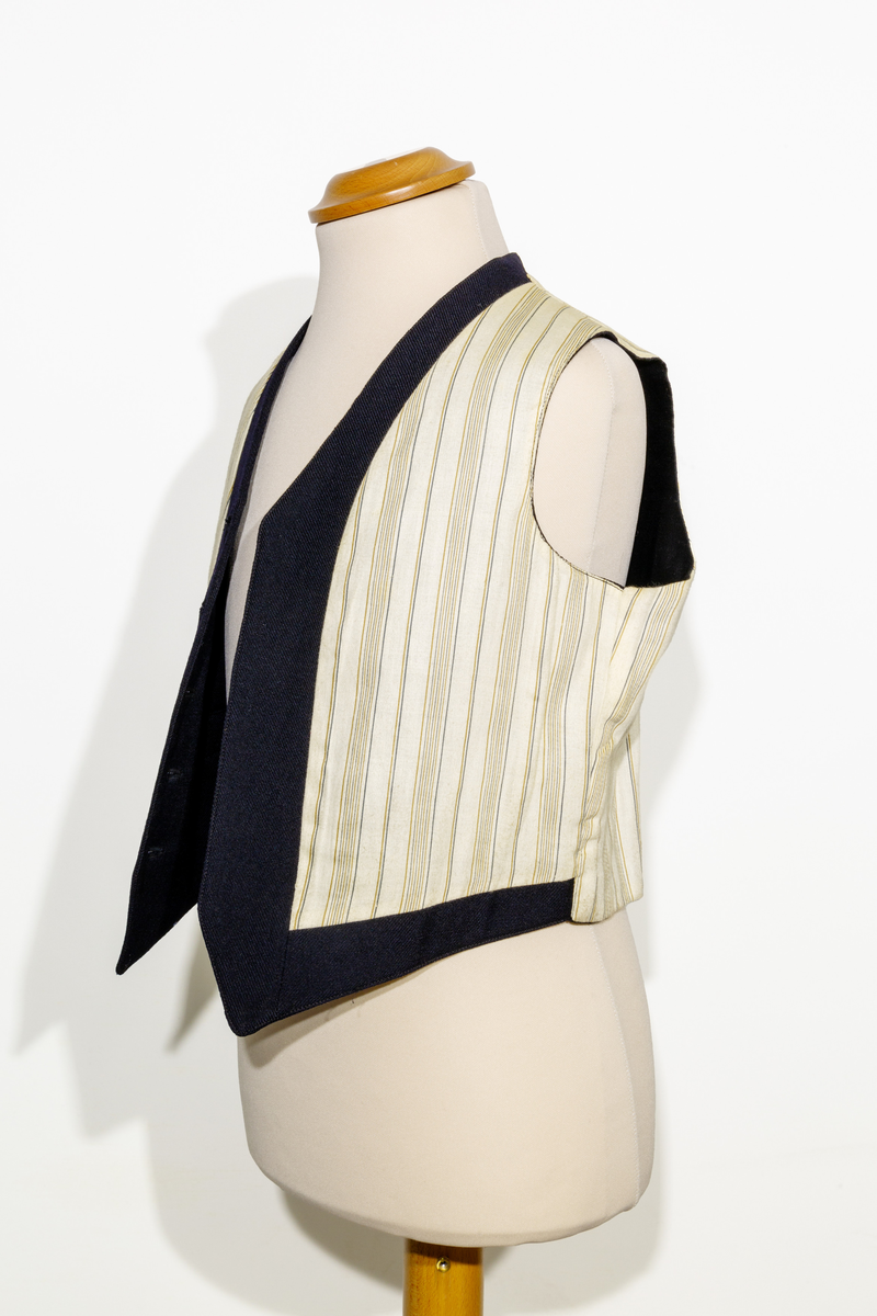 Uniformsväst för barn i mörkblått ylletyg. Västen har två fickor, en på vardera sidan om den främre knäppningen. Västen har fem stycken gulfärgade knappar med motiv av ankare. Västens bakstycke är i svart ylletyg och försedd med ett spänne. Västen är fodrad med ett randigt tyg med bruna och blå ränder på oblekt bas. 