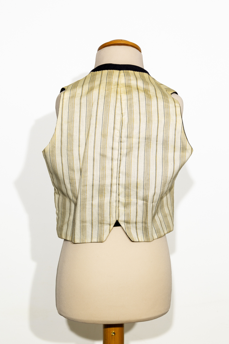 Uniformsväst för barn i mörkblått ylletyg. Västen har två fickor, en på vardera sidan om den främre knäppningen. Västen har fem stycken gulfärgade knappar med motiv av ankare. Västens bakstycke är i svart ylletyg och försedd med ett spänne. Västen är fodrad med ett randigt tyg med bruna och blå ränder på oblekt bas. 