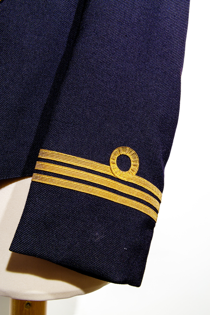 Uniformsjacka (marinen) till barn i mörkblått ylletyg. Försedd med fem rader av guldfärgade knappar med motiv av ankare. Ärmarna pryds av tre rader med guldfärgat band. Jackan är fodrar med ett svart tyg och ett randigt tyg med blå och bruna ränder på oblekt bas på ärmarnas insida. Jackan har en inneficka på höger sida. I jackans nackstycke finns en etikett med texten "Barngarderoben Falun" samt en hängare. 