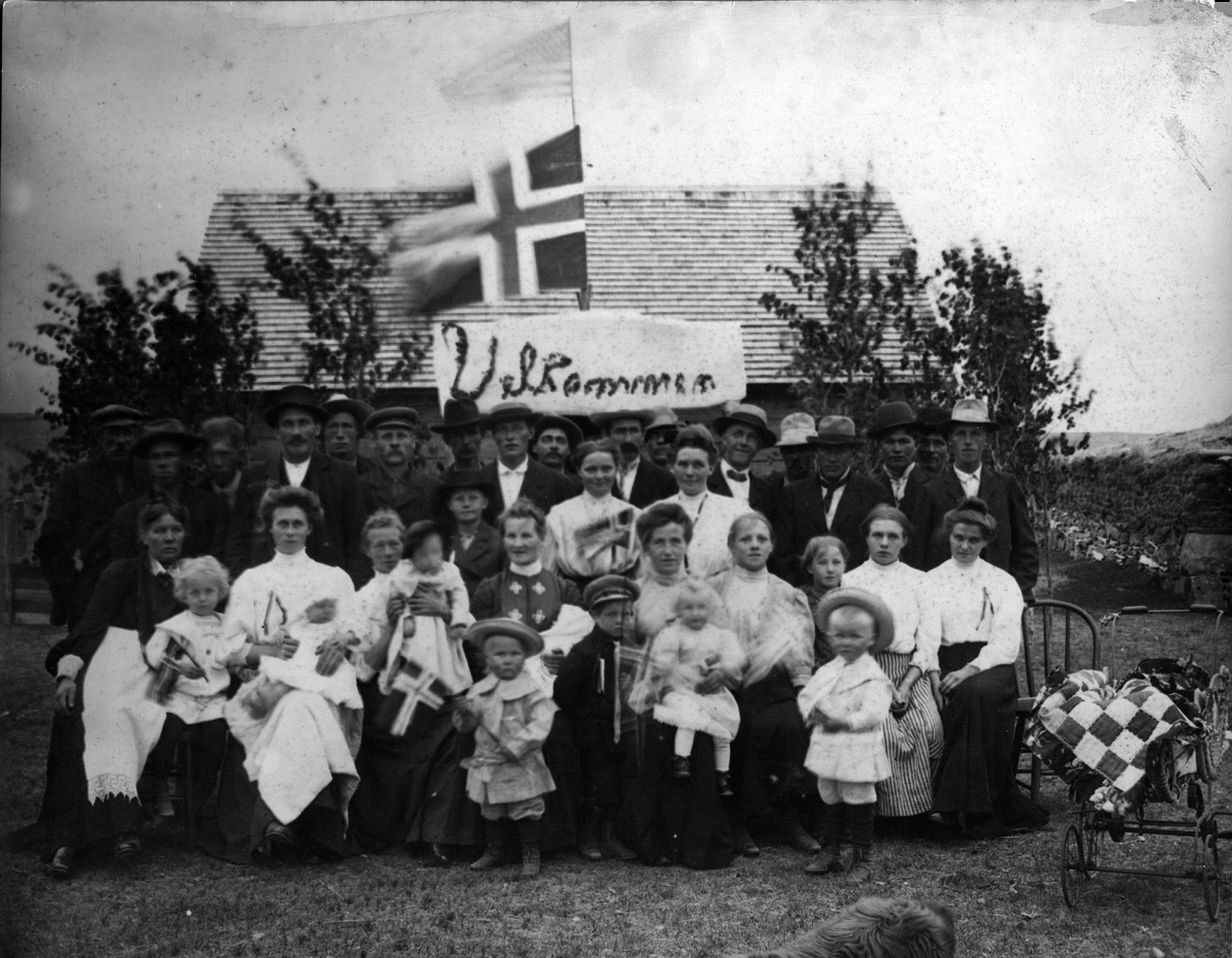 Stort  gruppebilde av mennesker med det norske og det amerikanske flagg og en plakat som sier "Velkommen" i bakgrunnen. Skrevet på baksiden av bildet: J. J. Gilseth, Mohall, N. Dak.