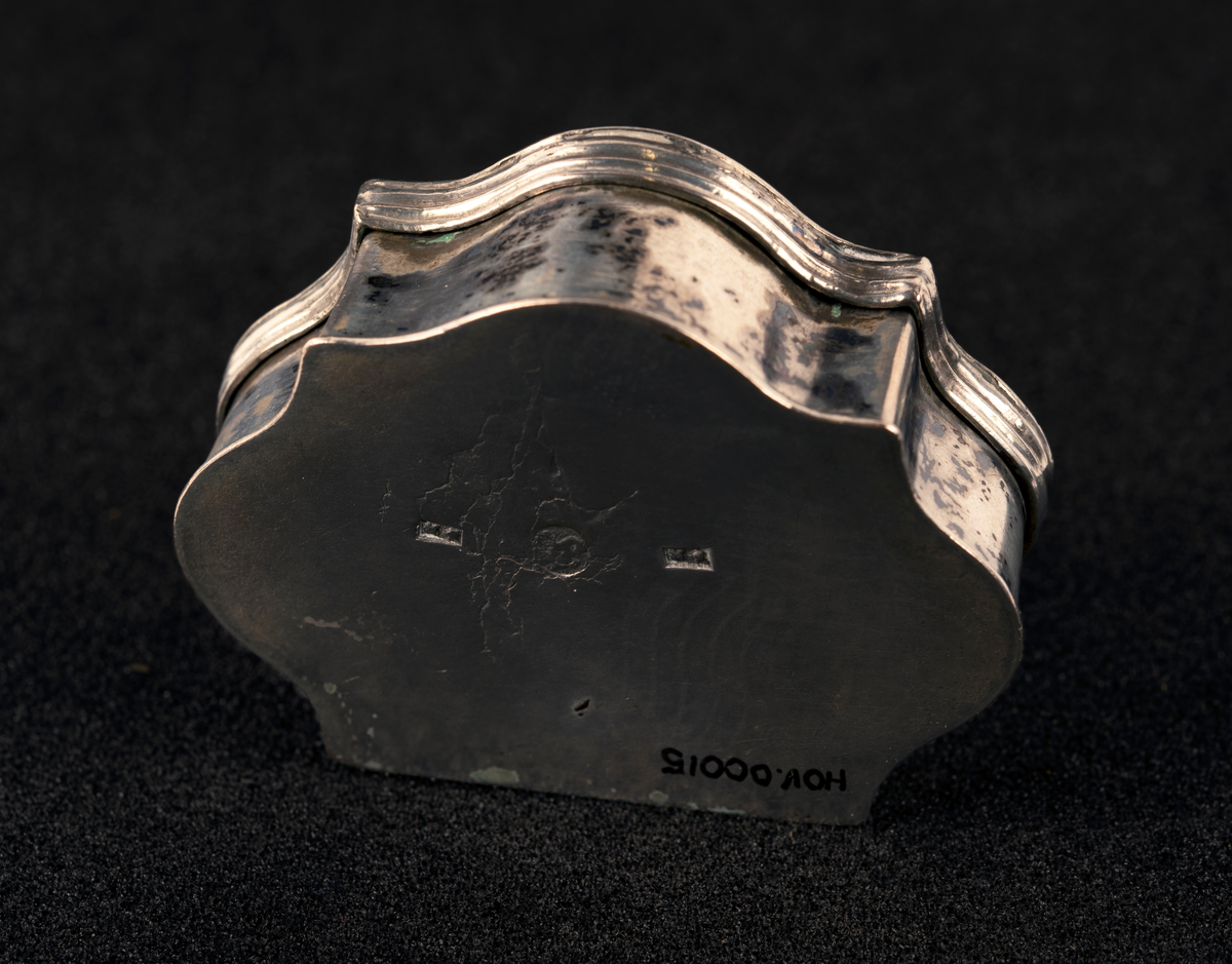 Dåse av sølv, med buede kanter og hengslet lokk med profilert kant. Gravert borddekor rundt initialer på lokket, utydelig stempel.