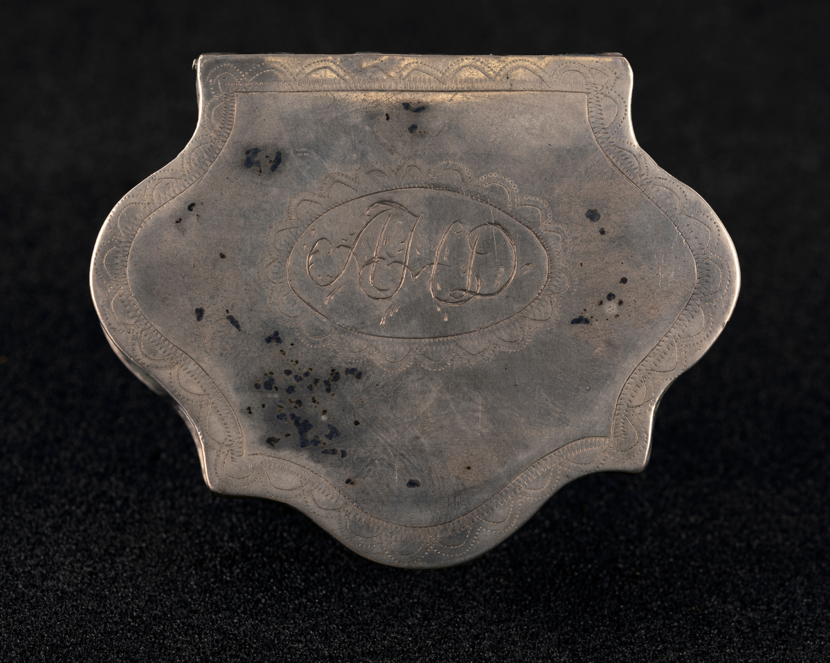 Dåse av sølv, med buede kanter og hengslet lokk med profilert kant. Gravert borddekor rundt initialer på lokket, utydelig stempel.
