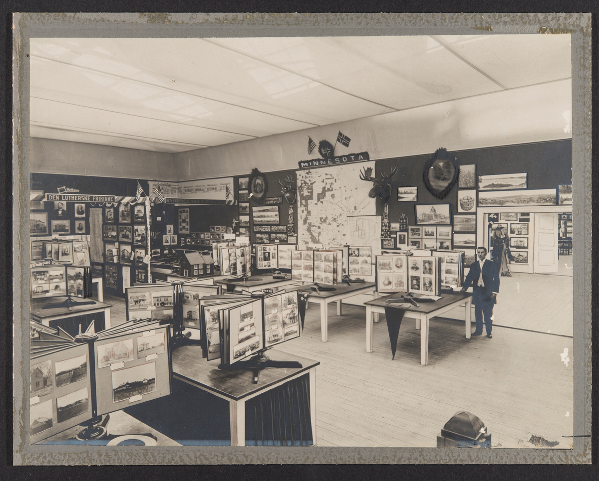 Oslo, 1914.
Utstillingssal med fotografier m.m.
Tekst bl.a. "Minnesota"
"Det utflyttede Norges utstilling 1914.
Utstillingssalen".