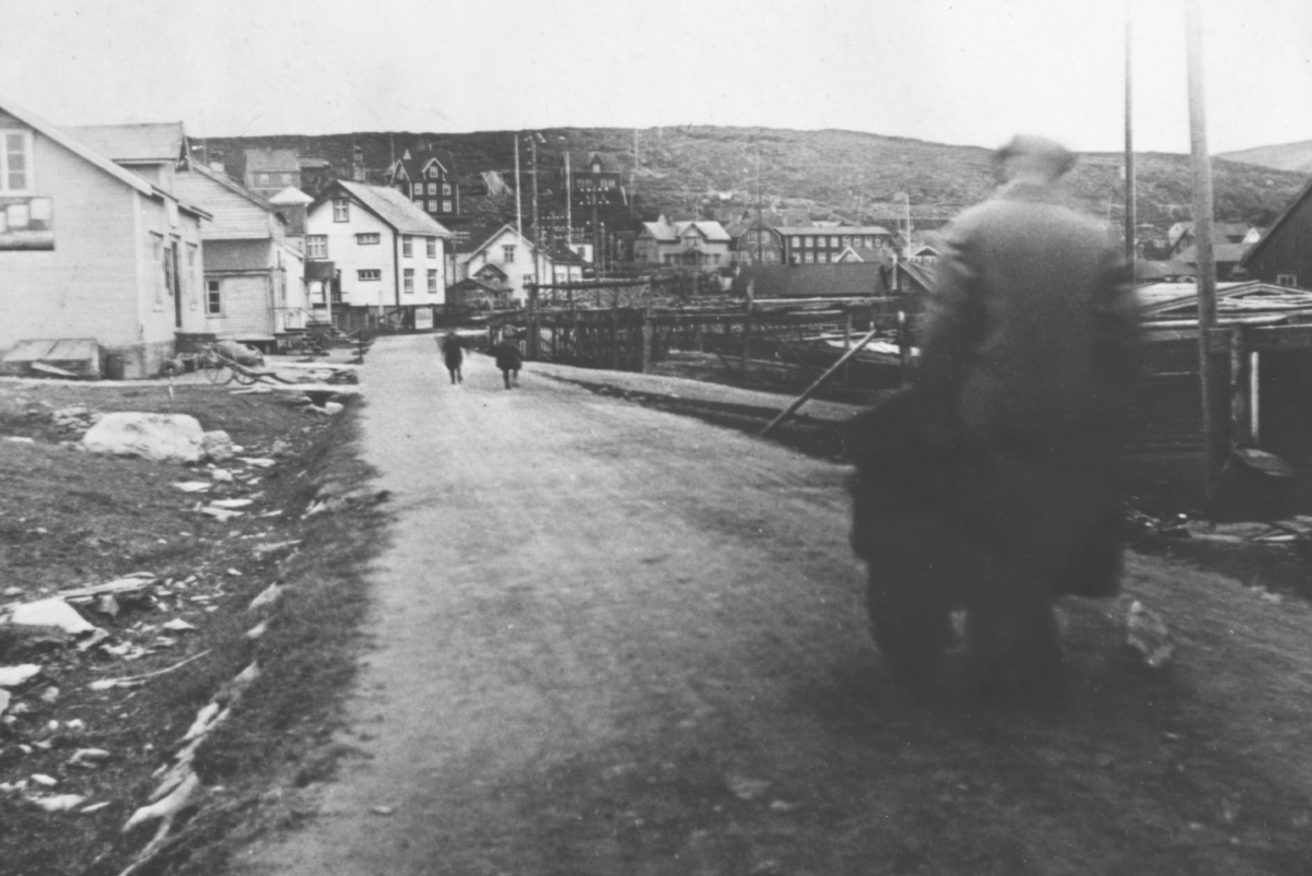 Mehamn, 1938. Bygninger, vei med to barn, en mann med ryggen til, fiskehjeller ned mot havna til høyre for veien.