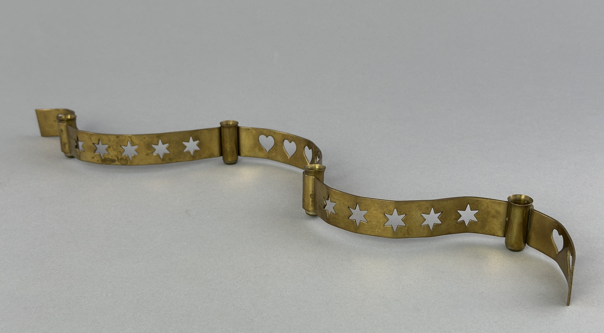 Ljusstake, "Slingan" för fyra ljus, består av ett mässingband som böjts i mjuk vågform och dekorerats med stansade stjärnor och hjärtan i grupper om fem. Mellan dessa grupper sitter ljushållare fastlödda med bandet.