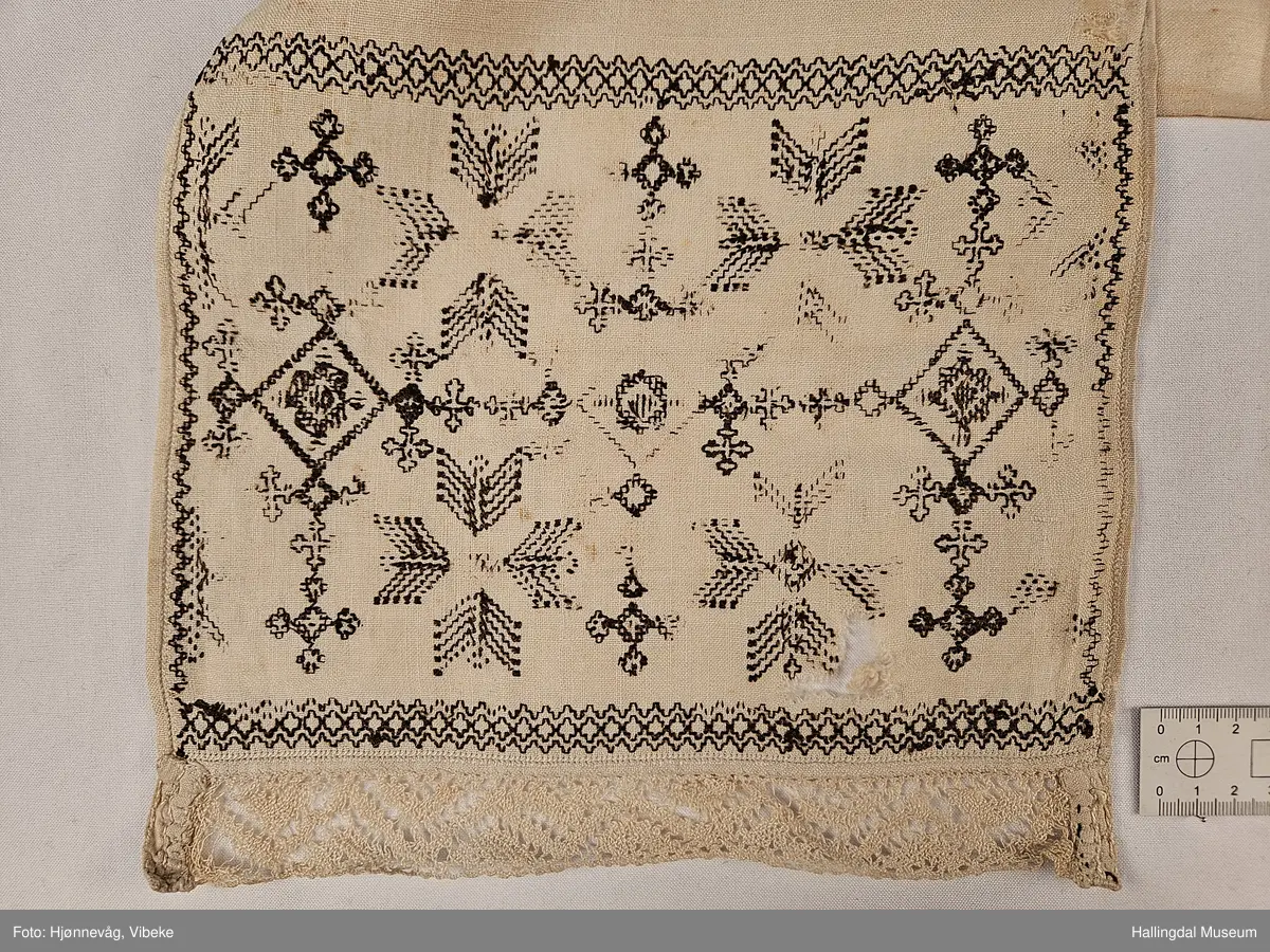 Lin, sauma, sydd. Sydd av 3 rektangulære stykke linty, i midten eit som er 54,0 x 54,0 cm, i kvar ende eit som er 29,5 x 26,8 cm. Svartsaum i ei lengde av 45 cm og br.: 2,7 cm langs eine langsida på midtstkket. Ytst på endestykka svartsaum i ei lengde av 19,2 cm over heile bredda. 3,2 cm breid blondekant langs båe stuttendane. 