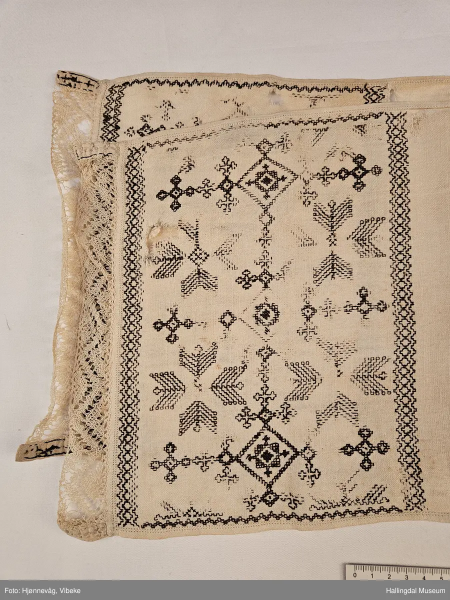Lin, sauma, sydd. Sydd av 3 rektangulære stykke linty, i midten eit som er 54,0 x 54,0 cm, i kvar ende eit som er 29,5 x 26,8 cm. Svartsaum i ei lengde av 45 cm og br.: 2,7 cm langs eine langsida på midtstkket. Ytst på endestykka svartsaum i ei lengde av 19,2 cm over heile bredda. 3,2 cm breid blondekant langs båe stuttendane. 