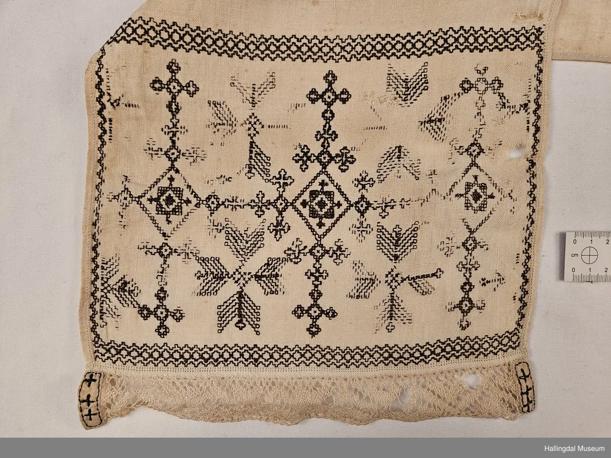 Lin, sauma, sydd. Sydd av 3 rektangulære stykke linty, i midten eit som er 54,0 x 54,0 cm, i kvar ende eit som er 29,5 x 26,8 cm. Svartsaum i ei lengde av 45 cm og br.: 2,7 cm langs eine langsida på midtstkket. Ytst på endestykka svartsaum i ei lengde av 19,2 cm over heile bredda. 3,2 cm breid blondekant langs båe stuttendane. 