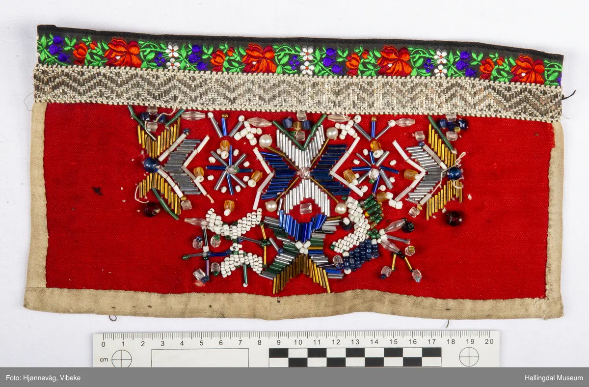 Ullstoff, bomullstoff, bånd, perler og kårilsteiner. Firkanta , raudt ullstoff, fora med kvitt bomullstoff.
Boler øvst, dekor med perler og kårilstein. Kanta med boler øvst, sauma med perler og kårilstein i 8-bladrøser.
