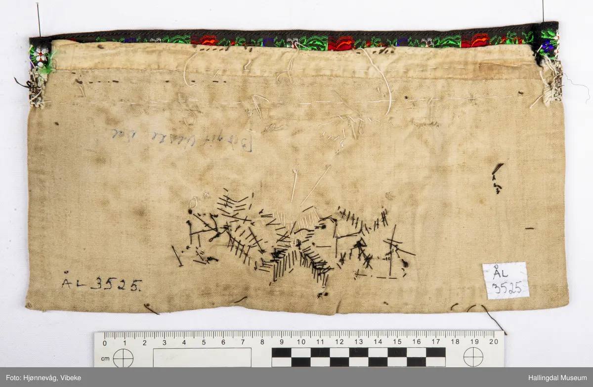 Ullstoff, bomullstoff, bånd, perler og kårilsteiner. Firkanta , raudt ullstoff, fora med kvitt bomullstoff.
Boler øvst, dekor med perler og kårilstein. Kanta med boler øvst, sauma med perler og kårilstein i 8-bladrøser.