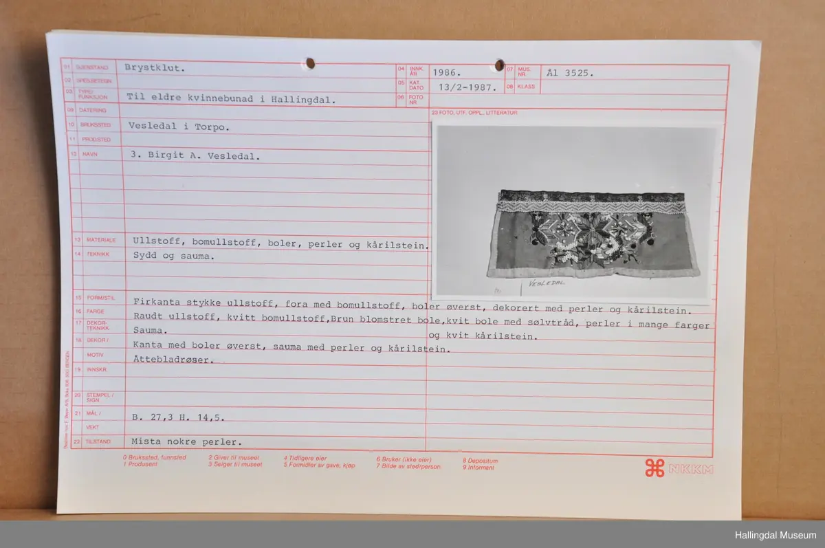 Ullstoff, bomullstoff, bånd, perler og kårilsteiner. Firkanta , raudt ullstoff, fora med kvitt bomullstoff.
Boler øvst, dekor med perler og kårilstein. Kanta med boler øvst, sauma med perler og kårilstein i 8-bladrøser.