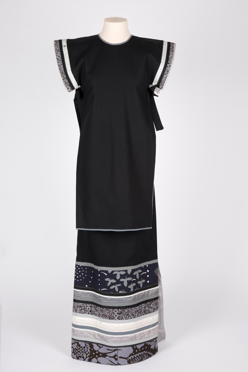 Todelt selskapskjole. Maskinsydd av to-skafts svart bomullsstoff, helfôret med grå syntetisk silke. Lang "Herold"-jakke- overdel, åpen i sidene og med smalt belte. Split i nakken med hekte. Overdelen er uten ermer, men med båndapplikasjoner rundt armringningen av finske stoffer i grått, mørkt brunt og hvitt. Rett skjørt, ankelsidt med høye splitter i sidene, nedre del med tverrgående applikasjoner av samme finske stoffer.