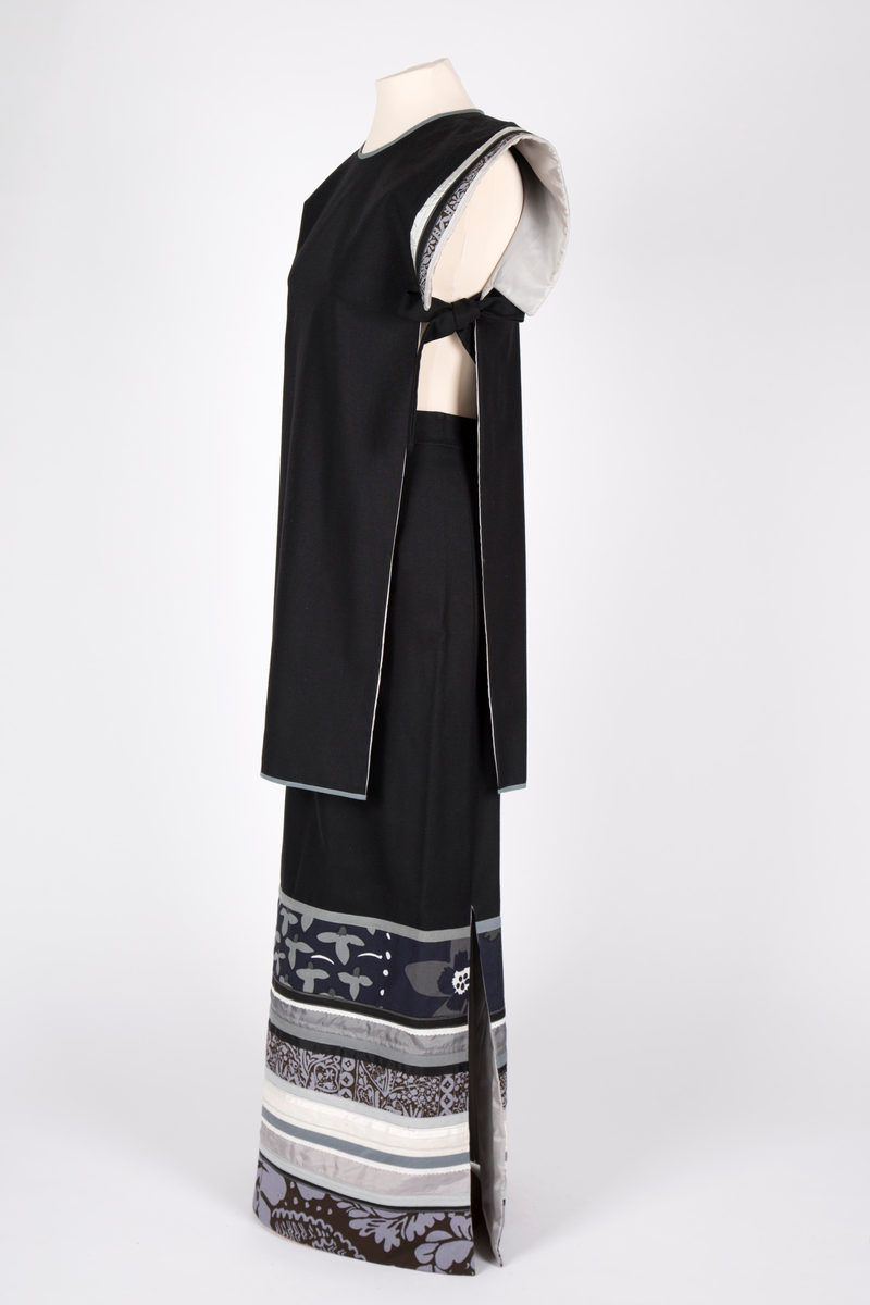 Todelt selskapskjole. Maskinsydd av to-skafts svart bomullsstoff, helfôret med grå syntetisk silke. Lang "Herold"-jakke- overdel, åpen i sidene og med smalt belte. Split i nakken med hekte. Overdelen er uten ermer, men med båndapplikasjoner rundt armringningen av finske stoffer i grått, mørkt brunt og hvitt. Rett skjørt, ankelsidt med høye splitter i sidene, nedre del med tverrgående applikasjoner av samme finske stoffer.