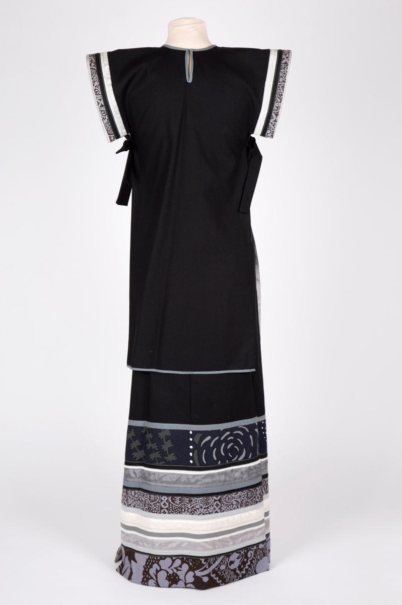 Todelt selskapskjole. Maskinsydd av to-skafts svart bomullsstoff, helfôret med grå syntetisk silke. Lang "Herold"-jakke- overdel, åpen i sidene og med smalt belte. Split i nakken med hekte. Overdelen er uten ermer, men med båndapplikasjoner rundt armringningen av finske stoffer i grått, mørkt brunt og hvitt. Rett skjørt, ankelsidt med høye splitter i sidene, nedre del med tverrgående applikasjoner av samme finske stoffer.