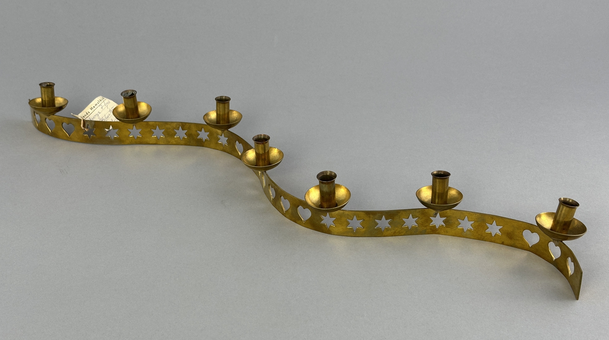 Ljusstake, "Ljusslingan" för sju ljus, består av ett mässingband som böjts i mjuk vågform och dekorerats med stansade stjärnor och hjärtan. Ljushållarna är fastlödda på bandets högkant.