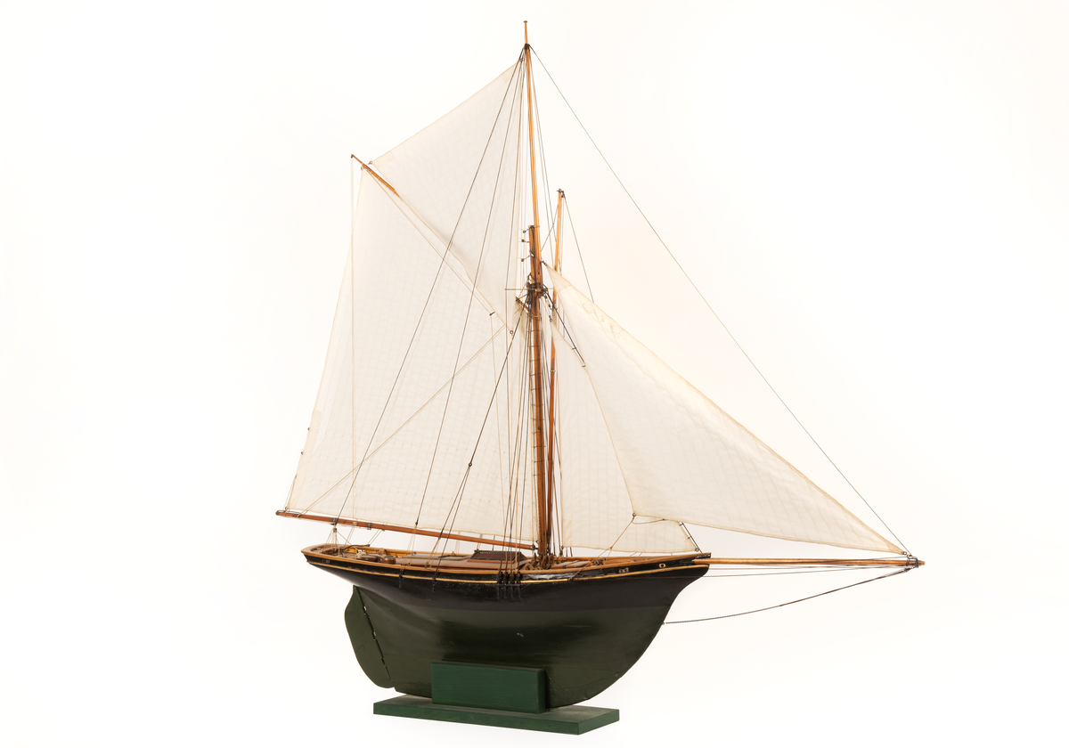 Båtmodell av segelkuttern HELMI, byggd i ett stycke med däcksinredning oc överhängande stäv. Akterspegel med hackbräde. Roder med rorkult av järn. Riggad, mast med stång, gaffelsegel, fock och klyvare. Svartmålad, förgylld list, grön vattengång. Fernissat däck. Tillhörande monter: svart.