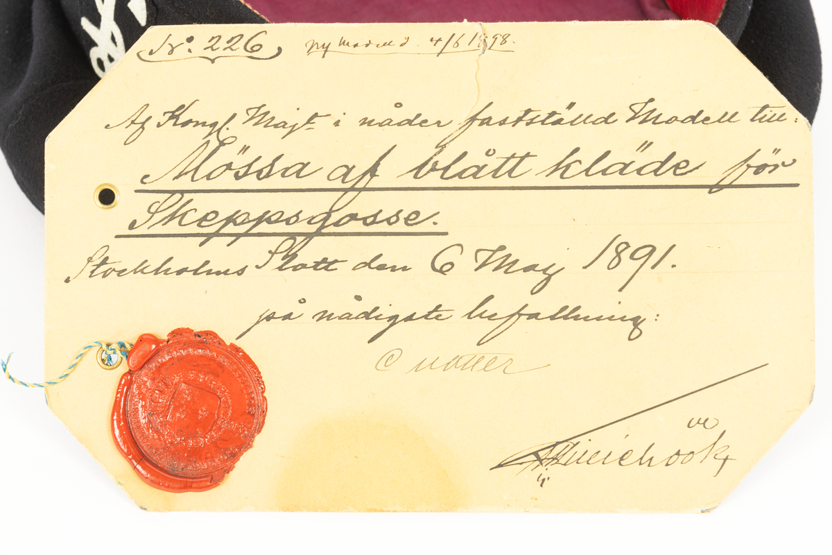 Rund mössa av blått kläde för skeppsgosse M/ 1891. Lila foder och röd svettrem.