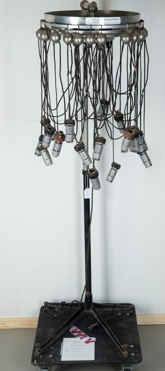 Skriv om texten
sittande på arm av metall som skjuter ut från  ett äggformat hölje av plåt inom vilken torkapparatens motor sitter. På toppen en dekorativ formation av blått glas på vilken det står instansat. "Wella" samt fabrikatets logotyp i form av ett kvinnohuvud med långt hår. På utsidan bockade stålrör som vilka man kan ta tag i som handtag när man vill förflytta torken. På ena sidan sitter ena änden av en elektrisk kabel av vit plast i vars andra ände en stickkontakt av svart plast sitter. Vid användning av apparaten sticks denna i vägguttag så att ström tillförs och maskinen kan användas. Det hela sitter på en ställing bestående av vertikalt stående stålrör av metall med fyrarmad fot med hjul så att ställningen kan förflyttas lätt. Stålröret är tvådelat med den ena delen instucken i den andra och med hjälp av en skruv av plast kan ställas i olika höjd. Metallskylt med text: Wella Darmstadt" samt upplysningar för typ, nr, strömstyrka. På plasthuvens utsida en något böjd och deformerad skylt i form av utskurna silverfärgade bokstäver i plast: "Wellapot". Placeras ovanför en sittande persons huvud för att torka håret med.