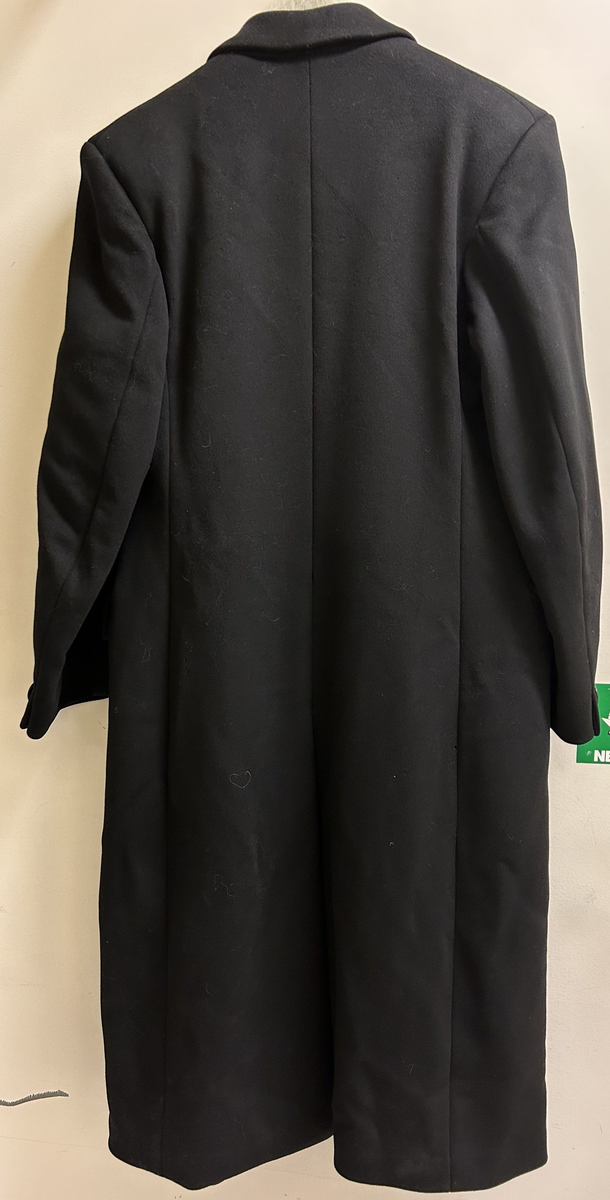 Mansrock i svart ylletyg. Rak modell med liggande krage. Två fickor med lock. Dubbelknäppning med tre par knappar. Mattlaserad svart satinvävt foder. Två innerfickor i passpoal. Rak ärm med knapp. Figursytt bakstycke.
Ett hål nedtill på vänster insida ("avigsidan").