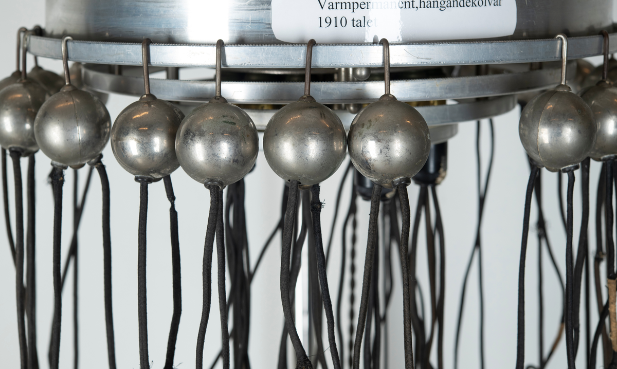 Skriv om texten
sittande på arm av metall som skjuter ut från  ett äggformat hölje av plåt inom vilken torkapparatens motor sitter. På toppen en dekorativ formation av blått glas på vilken det står instansat. "Wella" samt fabrikatets logotyp i form av ett kvinnohuvud med långt hår. På utsidan bockade stålrör som vilka man kan ta tag i som handtag när man vill förflytta torken. På ena sidan sitter ena änden av en elektrisk kabel av vit plast i vars andra ände en stickkontakt av svart plast sitter. Vid användning av apparaten sticks denna i vägguttag så att ström tillförs och maskinen kan användas. Det hela sitter på en ställing bestående av vertikalt stående stålrör av metall med fyrarmad fot med hjul så att ställningen kan förflyttas lätt. Stålröret är tvådelat med den ena delen instucken i den andra och med hjälp av en skruv av plast kan ställas i olika höjd. Metallskylt med text: Wella Darmstadt" samt upplysningar för typ, nr, strömstyrka. På plasthuvens utsida en något böjd och deformerad skylt i form av utskurna silverfärgade bokstäver i plast: "Wellapot". Placeras ovanför en sittande persons huvud för att torka håret med.