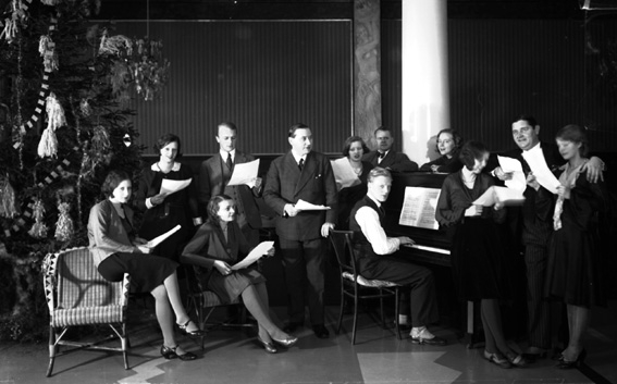 Man sitter vid piano, runt honom sitter det både män och kvinnor med noter. 
Fotografens ant: Fru Greta Bergsson.