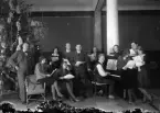 Man sitter vid piano, runt honom sitter det både män och kvinnor med noter. 
Fotografens ant:Fru Greta Bergsson.