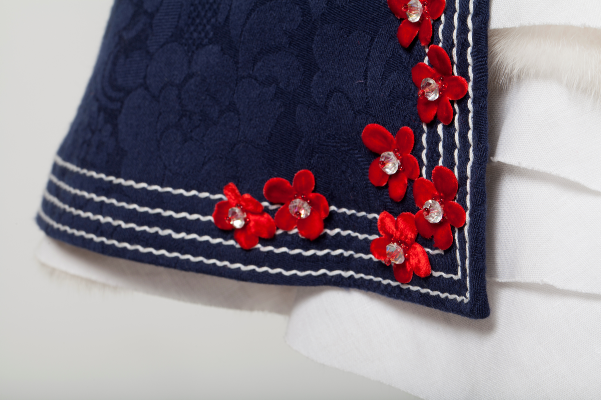 Frontal smekke med sentralt plassert, brodert tre eller grener med påsydde småblomster. Parallelle stikninger i kontrasterende farge, ispedd tilsvarende småblomster.