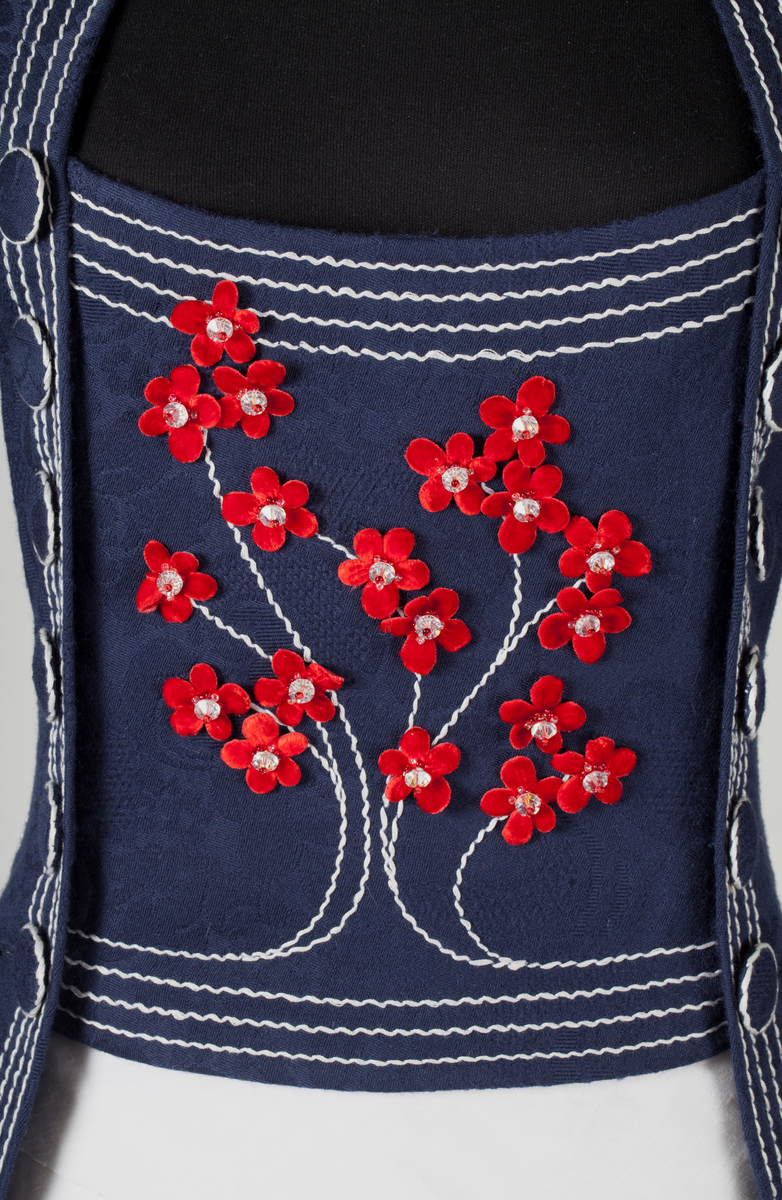 Frontal smekke med sentralt plassert, brodert tre eller grener med påsydde småblomster. Parallelle stikninger i kontrasterende farge, ispedd tilsvarende småblomster.
