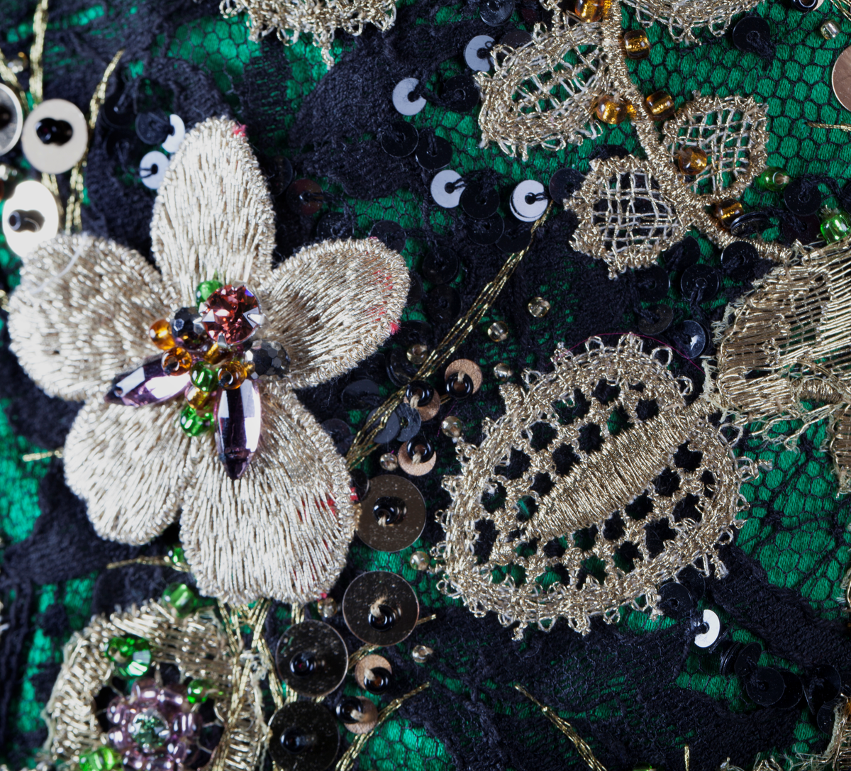 Småblomster og blader som vokser opp av en blomstereng, alt av gull, mot en bakgrunn av storblomstret tyllblonde.