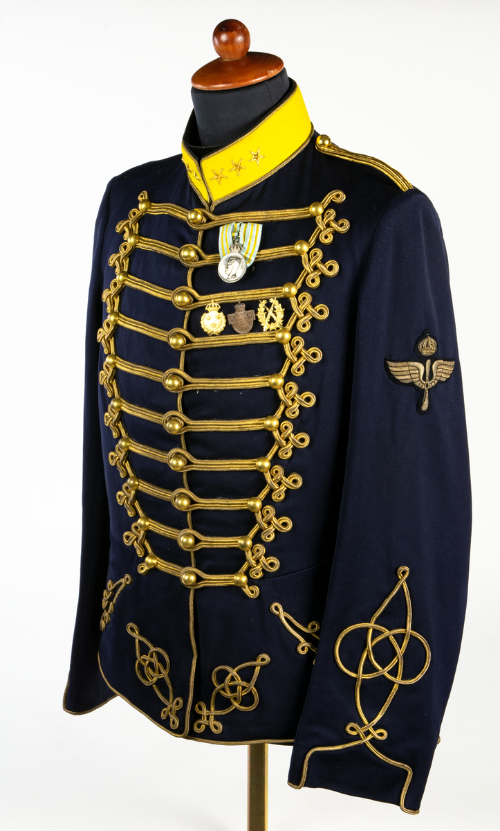 Dolma m/1895. K 4, Smålands husarregemente (gul krage), tre stjärnor på kragen för överste och flygemblem på vänster överarm. På bröstets vänstra: Gustav V:s olympiska medalj 1912. Fältidrottsmärke i guld. Riksidrottsmärke i brons nr 150310. Skyttemärke i guld för frivilliga skytteföreningar.