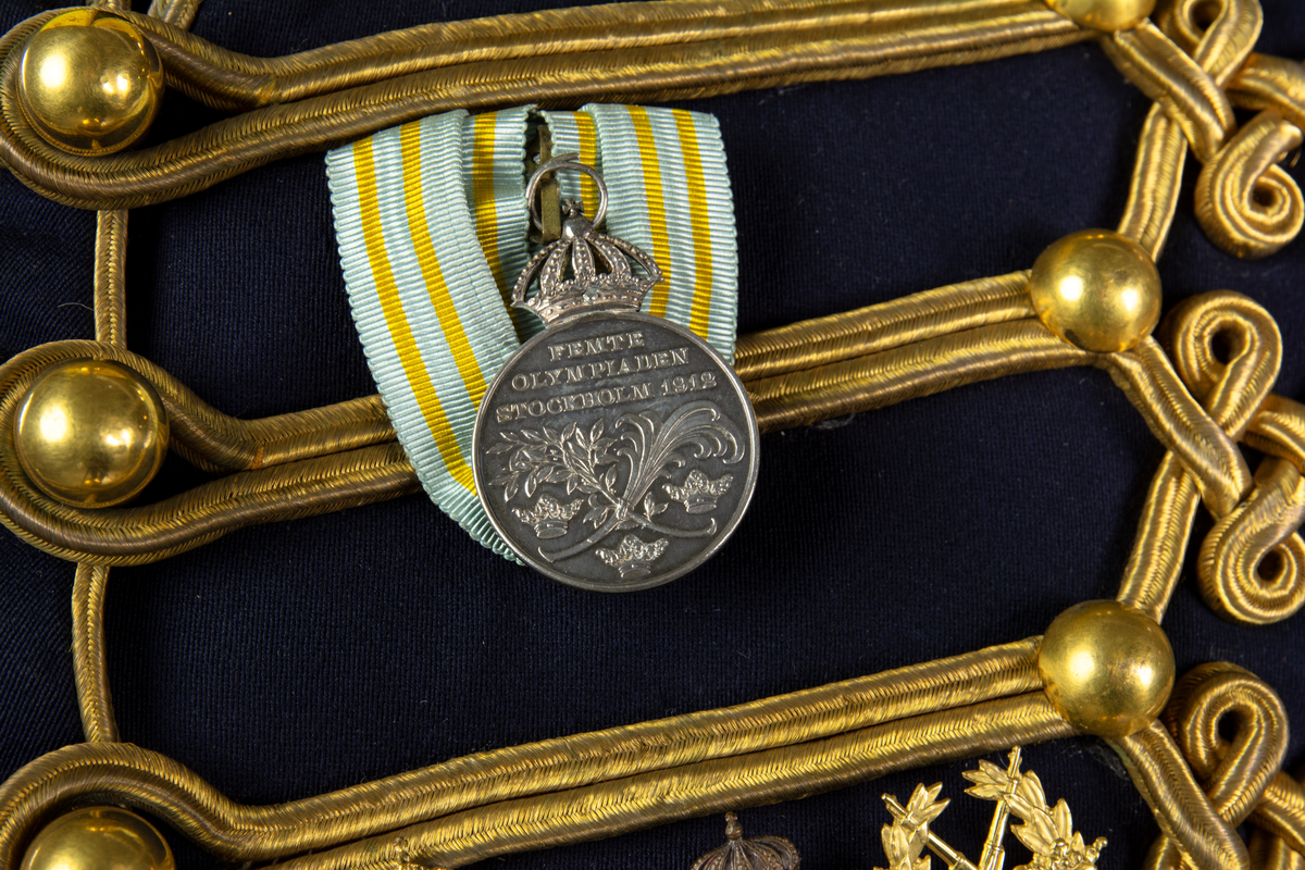 Dolma m/1895. K 4, Smålands husarregemente (gul krage), tre stjärnor på kragen för överste och flygemblem på vänster överarm. På bröstets vänstra: Gustav V:s olympiska medalj 1912. Fältidrottsmärke i guld. Riksidrottsmärke i brons nr 150310. Skyttemärke i guld för frivilliga skytteföreningar.