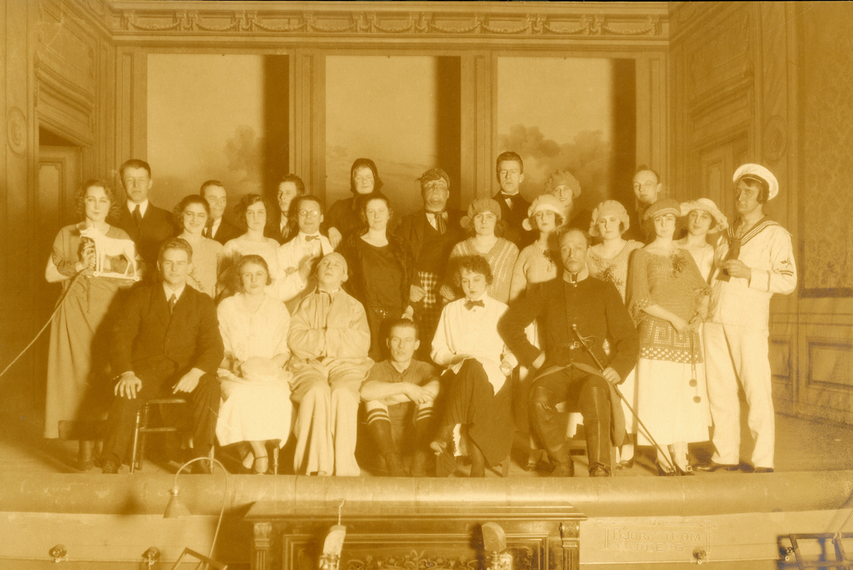 Ensemblen i nyårsrevyn "Än leve de gamla gudar!" 1924 på Varbergs teater. Efter premiären den 1 januari fick revyn först ett ljumt mottagande i recensionen av "Brutus" i Varbergsposten: "Anrättningen befanns vara skäligen mager...". Men efter viss omarbetning och mer repetitioner till föreställningen en vecka senare fick den idel lovord. Bland artisterna fanns kuplettsångarna Ernst Dahlqvist och herr Rosengren. Komikern B G Möllerström fick enligt Brutus publiken att tjuta av förtjustning åt roller som "lantliga Truls", slaktardräng, beväring och kapellmästare. Den verklige kapellmästaren hette Dahlgren och fick beröm, liksom "Jassflickorna" och "Valkyriorna". Men allra mest imponerade M Aschjem som både tränat in baletterna och målat dekorerna: "Aldrig förut har en lokalrevy här kunnat ståta med så pampiga dekorationer och finfina dräkter." Här ses bl a en sjöman, en fotbollsspelare, en kvinna hållande en vargfigur på hjul i snöre och självaste Karl XII. Lokalrevyn gjordes till förmån för Varbergs Gymnastik- och Idrottsförening.
Bild 1: Tonat foto (F9358) med Björkströms logotyp monterat i omslag.
Bild 2: Glasplåt, nedanför scenen ses piano och notställ. 
(Se även bildnr GB2_2778-2785)