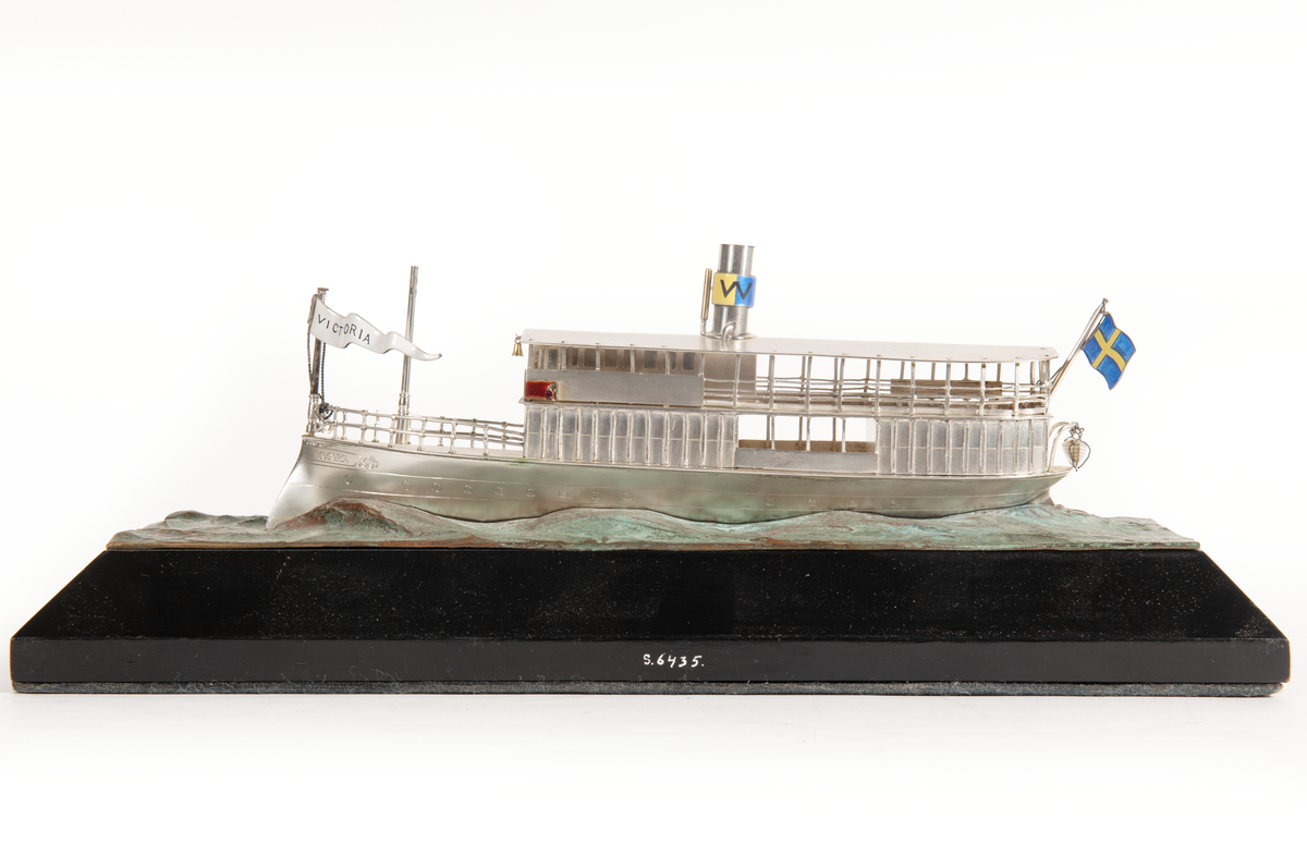 Fartygsmodell av  S/S VICTORIA (1882 vid Bergsund) i silver. Skrov med rammstäv, inbyggd brygga, skorsten med Waxholmsbolagets märke, ingraverat fartygsnamn på stäven, namnvimpel av silver, flagga akter. Livbåt akter. Monterad på vågimiterad kopparplatta och träsockel.
