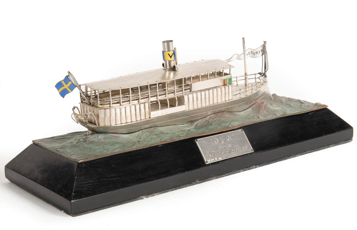 Fartygsmodell av  S/S VICTORIA (1882 vid Bergsund) i silver. Skrov med rammstäv, inbyggd brygga, skorsten med Waxholmsbolagets märke, ingraverat fartygsnamn på stäven, namnvimpel av silver, flagga akter. Livbåt akter. Monterad på vågimiterad kopparplatta och träsockel.