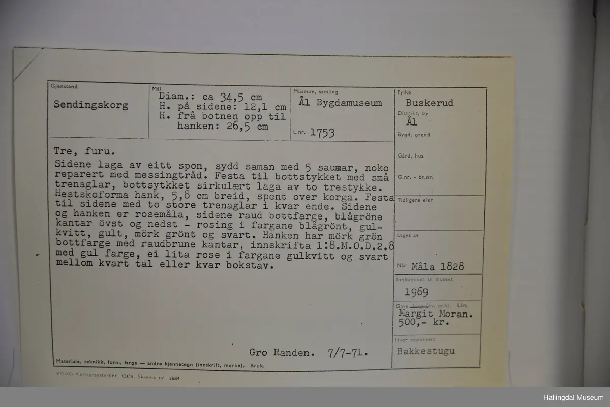 Sidene laga av eit spon, sydd saman av 5 saumar, noko reparert med messingtråd. Festa til bottstykket med små trenaglar. Bottstykket sirkulært laga av to trestykke. Hesteskoforma hank, spent over korga. Festa til sidene med to store trenaglar i kvar ende. Sidene og hanken er rosemåla, sidene i raud bottfarge, blågrøne kantar øvst og nedst - rosing i fargane blågrønt, gulkvitt, gult, mørk grønt og svart. Hanken har mørk grøn bottfarge med raudbrune kantar, innskrift med gul farge, ei lita rose i gulkvitt og svart mellom kvart tal eller bokstav.