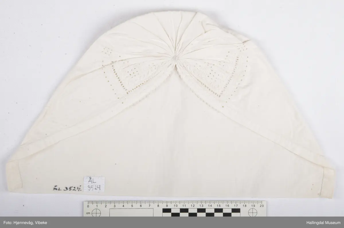 Bomullstoff og tråd. Sydd og brodert, hvitsøm. Rynket i nakken.