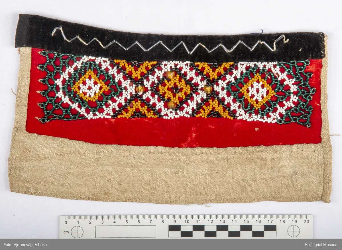 Brukt inni åpentrøye i Ålingsbunaden.  Klede, fløyelsbånd og perler. Perlenett. Sydd av rødt klede. Perler i svart, grønt, sennepsgult og kvitt. 4 store perler på midten. Kantet øverst med svart fløyelskant, 2,4 cm bredt. Sikksakkbånd av stavperler oppå fløyelskanten. Foret med linstoff i dreiel. Håndsydd. H: 14,5 cm, L: 26,5 cm.