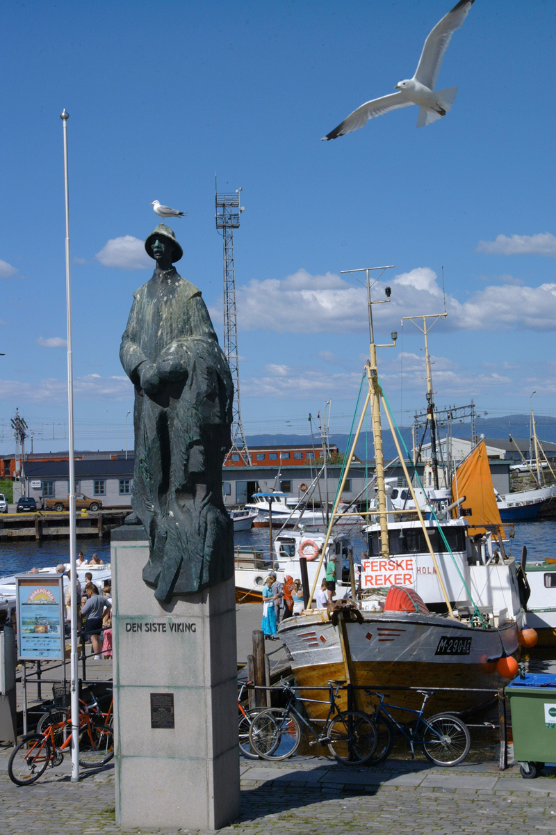Helfigur på sokkel. En fisker som står med sitt fiskegarn i favnen. Antrukket i lang arbeidsfrakk og sydvest på hodet.