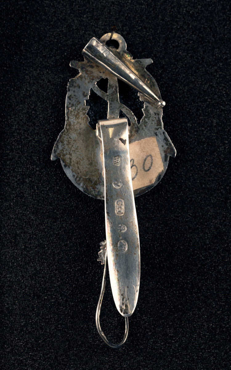 Nøstekrok av sølv til å hekte i beltet på damedrakt, formet som anker med blomsterkrans. Stempel: se foto.