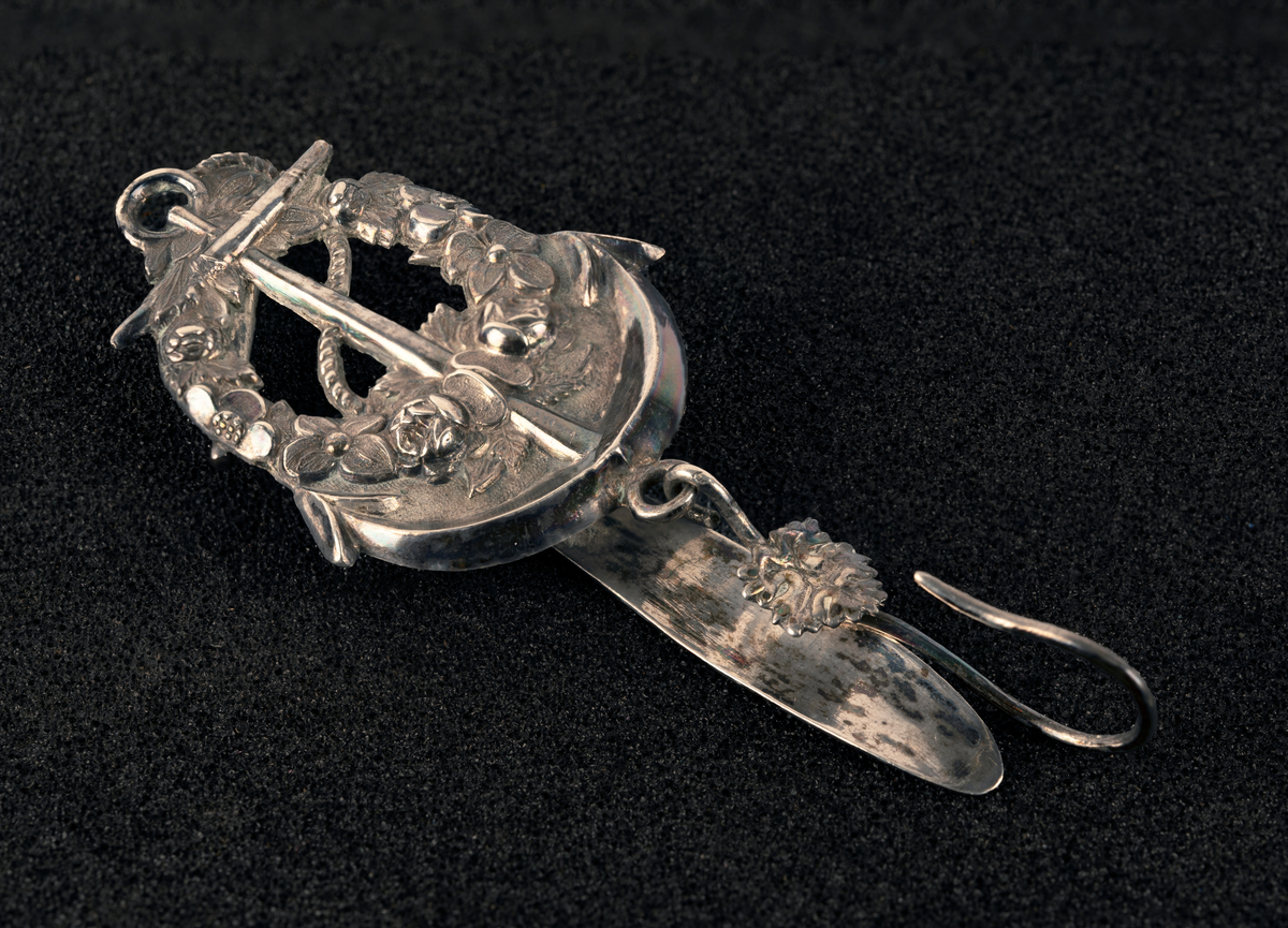 Nøstekrok av sølv til å hekte i beltet på damedrakt, formet som anker med blomsterkrans. Stempel: se foto.