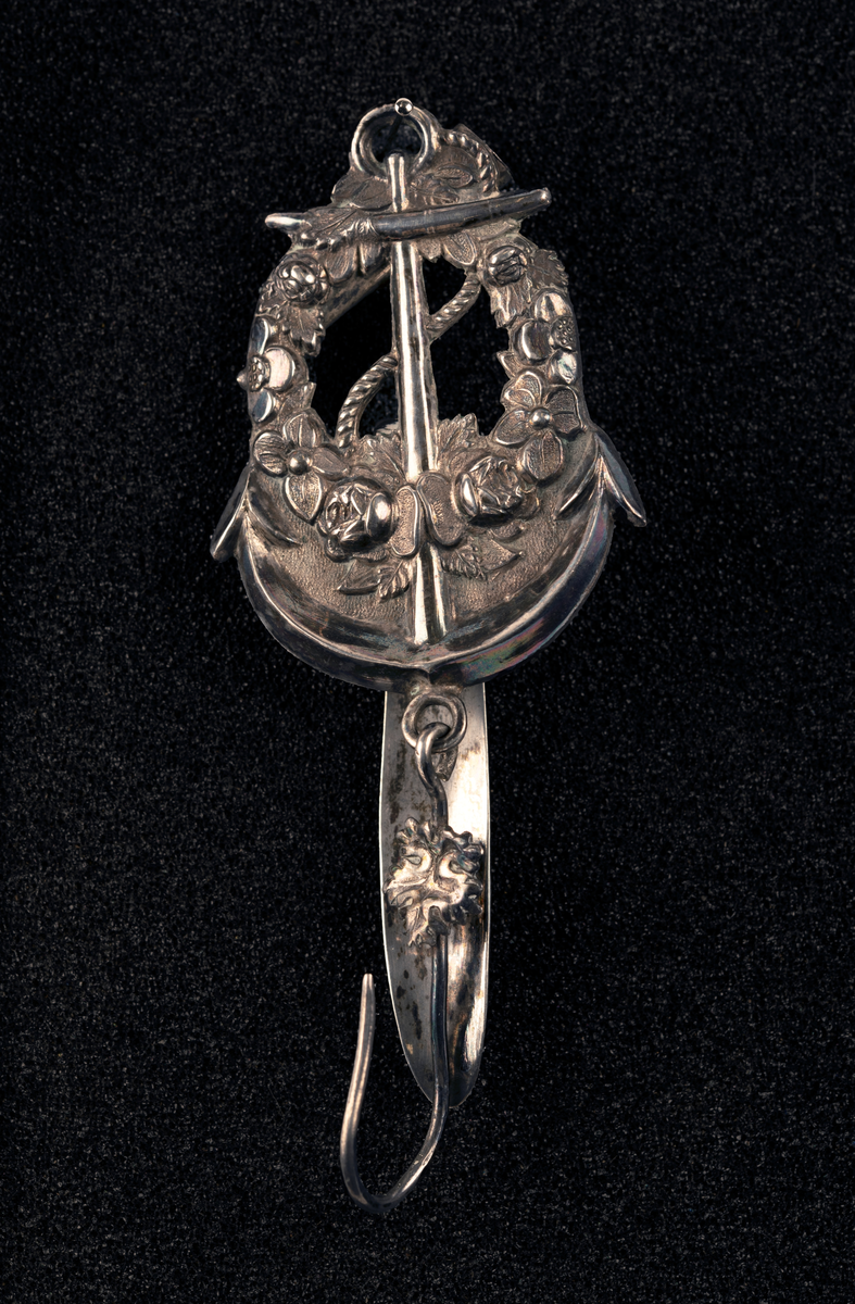 Nøstekrok av sølv til å hekte i beltet på damedrakt, formet som anker med blomsterkrans. Stempel: se foto.