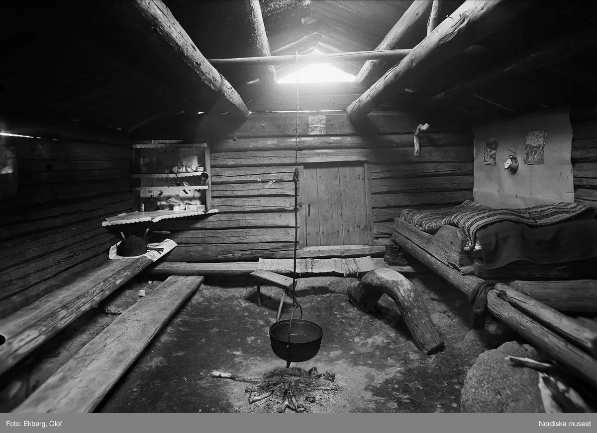 Eldhusinteriör. Juganes, Ärteråsens fäbod, Ore sn, Dalarna. Ett kokkärl hänger från taket ner över över eldstaden. Från taket silas ljuset ner. Längs väggarna väggfasta bänkar och säng. 