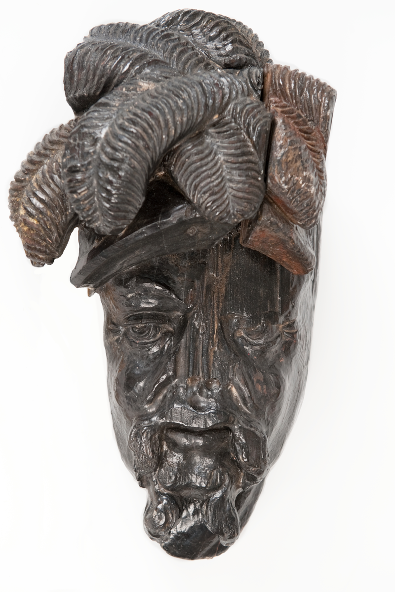 Del av skulptur. 
Huvudet till en högrest krigare med stor mustasch. På huvudet sitter hjälm med långa vajande plymer.