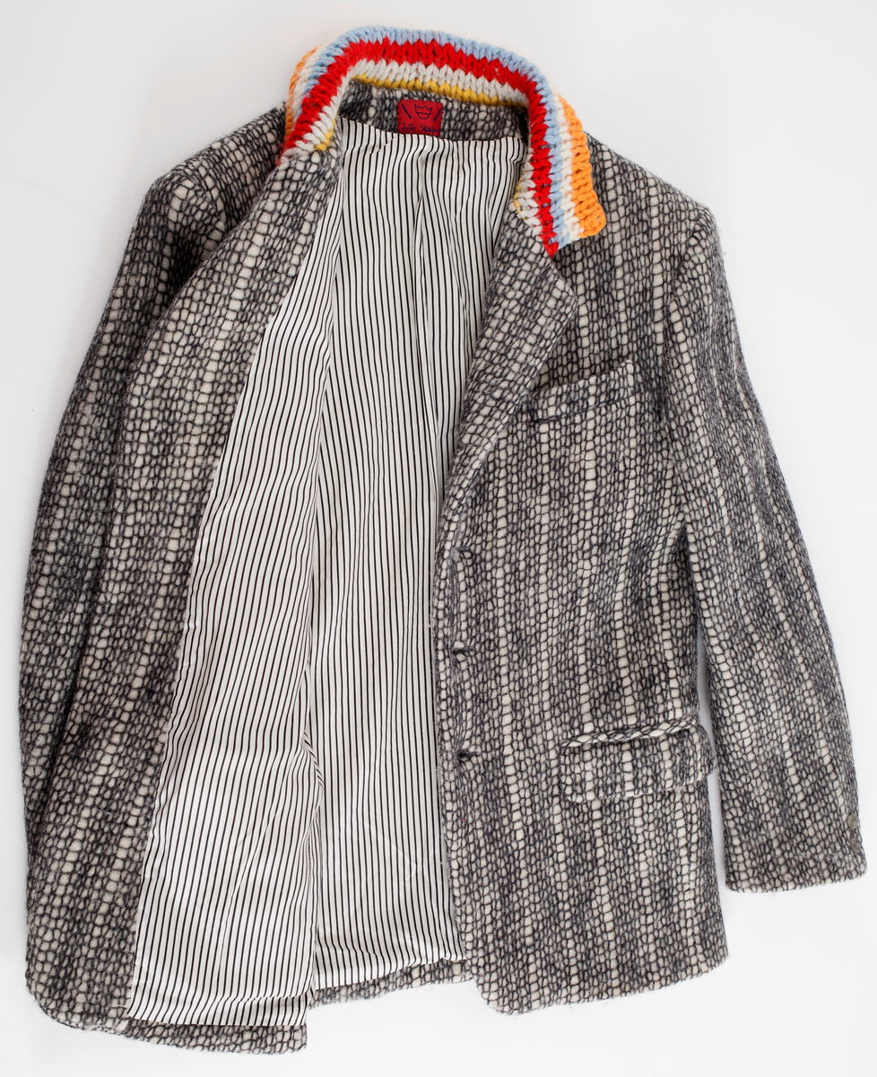 Dressjakke sydd av twool i koksgrå på hvit. Jakkens krage er strikket av tykt garn i ribbestrikk på tykke pinner i fargene (striper): oransje, gult, hvitt, lyseblått, rødt, gulbrunt og lysegrått.