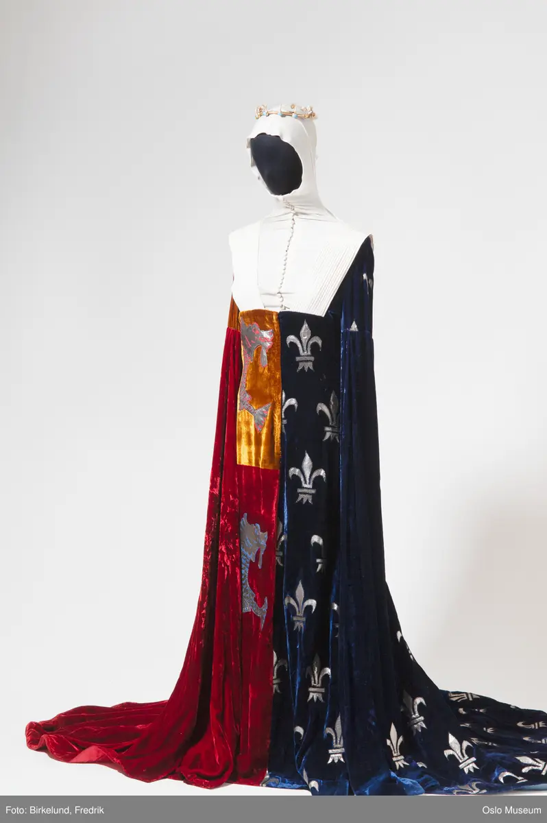 A= Kjole i rødbrun og blå fløyel med slep og lange ermer. Gullapplikasjoner med franske liljer og perler, applikasjoner i form av en drage. Hodeplagg, tettsittende hette i hvit silkelignende stoff med stretch og knapper.
B= Krone i metall med perler